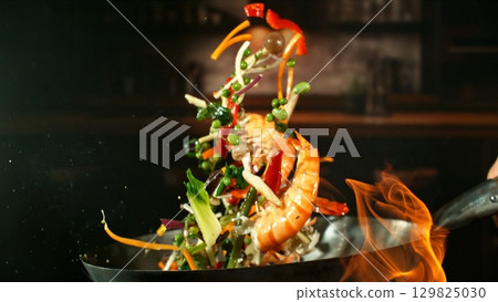 Freeze Motion of Wok Pan with Flying Ingredients in the Air and Fire Flames. Kitchen Interior on Background. Freeze Motion of Wok Pan with Flying Ingredients in the Air and Fire Flames. Kitchen Interior on Background. 129825030