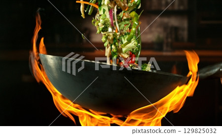 Freeze Motion of Wok Pan with Flying Vegetable Ingredients in the Air and Fire Flames. Kitchen Interior on Background. 129825032