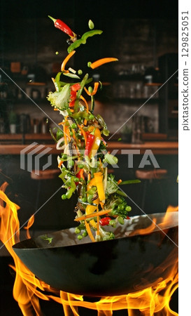 Freeze Motion of Wok Pan with Flying Vegetable Ingredients in the Air and Fire Flames. Kitchen Interior on Background. Vertical Aspect Ratio. 129825051