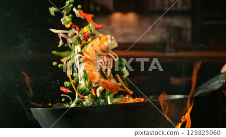 Freeze Motion of Wok Pan with Flying Ingredients in the Air and Fire Flames. Kitchen Interior on Background. 129825060