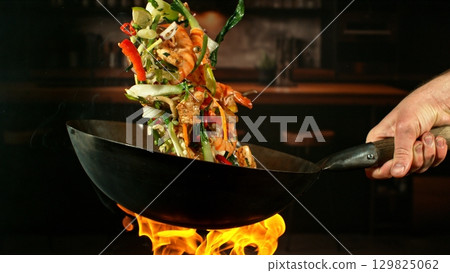 Freeze Motion of Wok Pan with Flying Ingredients in the Air and Fire Flames. Kitchen Interior on Background. 129825062