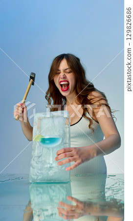 Woman smashing ice block with blue cocktail inside while laughing with dynamic energy 129826686