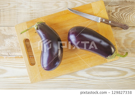 Fresh eggplants on cutting board and knife on wooden background, cooking fresh vegetables in kitchen, healthy food. Fresh eggplants on cutting board and knife on wooden background, cooking fresh vegetables in kitchen, healthy food. 129827338