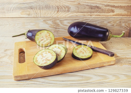 Fresh eggplants on cutting board and knife on wooden background, cooking fresh vegetables in kitchen, healthy food. Fresh eggplants on cutting board and knife on wooden background, cooking fresh vegetables in kitchen, healthy food. 129827339