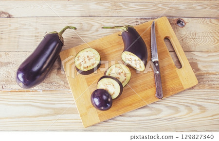 Fresh eggplants on cutting board and knife on wooden background, cooking fresh vegetables in kitchen, healthy food. Fresh eggplants on cutting board and knife on wooden background, cooking fresh vegetables in kitchen, healthy food. 129827340