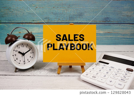 Sales Playbook text written on paper card with calculator and alarm clock on wooden background 129828043