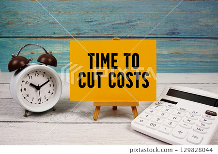 Time To Cut Costs text written on paper card with calculator and alarm clock on wooden background Time To Cut Costs text written on paper card with calculator and alarm clock on wooden background 129828044