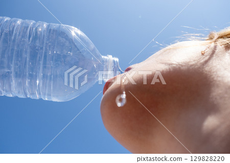 Young woman enjoying refreshing water from a plastic bottle under a bright blue summer sky, quenching her thirst on a hot day Young woman enjoying refreshing water from a plastic bottle under a bright blue summer sky, quenching her thirst on a hot day 129828220