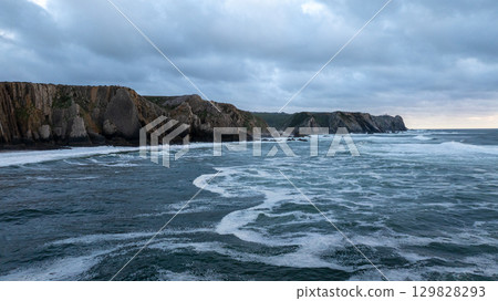 Dark, moody ocean waves crashing on the shore at dusk with cliffs in the background at Praia da Adraga beach, Sintra, Portugal 129828293