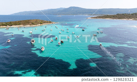 Numerous boats are anchored in the crystal clear turquoise waters of Vourvourou, Sithonia, Greece, surrounded by lush green islands, creating a picturesque summer scene Numerous boats are anchored in the crystal clear turquoise waters of Vourvourou, Sithonia, Greece, surrounded by lush green islands, creating a picturesque summer scene 129828321
