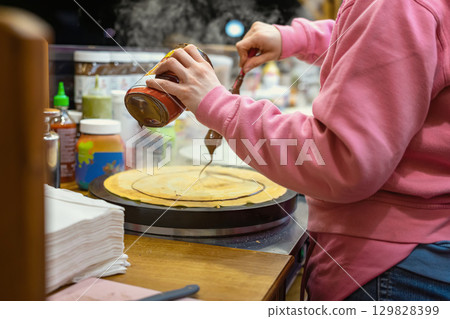 Chocolate crepe preparation at market. Woman pours chocolate on freshly cooked pancake at street food stall during winter market. Concept of sweet holiday food, festive culinary tradition 129828399