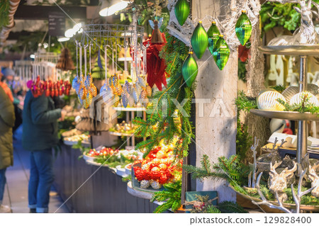 Christmas ornaments at festive market stall. Christmas market stall filled with shiny green and gold ornaments, pine decor, glowing lights. Concept handmade decorations and festive market charm Christmas ornaments at festive market stall. Christmas market stall filled with shiny green and gold ornaments, pine decor, glowing lights. Concept handmade decorations and festive market charm 129828400