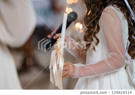 Young Flower Girl Holding Lit Candle at Orthodox Christian Wedding Ceremony, White Lace Dress, Soft Lighting 129828514