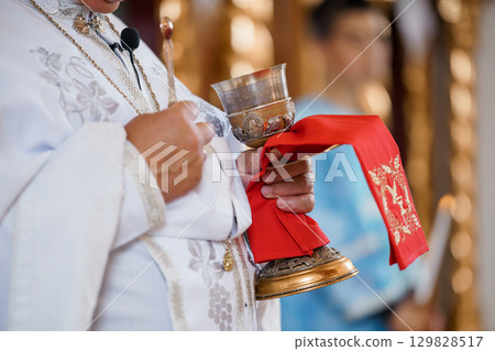 Eastern Orthodox Priest Holding Golden Chalice During Religious Ceremony, Red Cloth, White Vestments, Spiritual Ritual Photography. 129828517