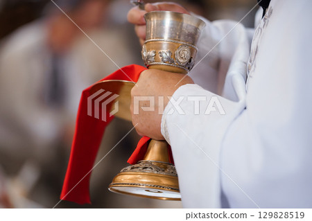 Solemn Catholic Ritual: Priest Holding Golden Chalice During Religious Ceremony, White Vestment, Red Sash, Sacred Communion, Golden Details. 129828519