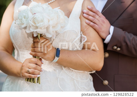 Intimate Wedding Embrace: Bride and Groom Holding Bouquet, White Peonies, Pearl Necklace, Brown Suit, Romantic Celebration, Close-Up View, Natural Lighting 129828522
