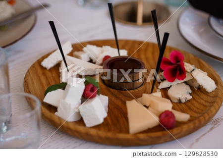 Gourmet Cheese Board with Central Dip, Raspberries, and Flowers on Wooden Surface, Brightly Lit Table Setting. 129828530