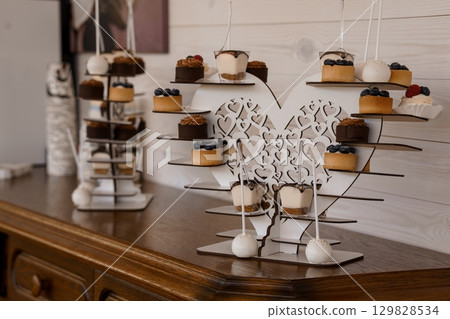 Elegant Wedding Dessert Display: Heart-Shaped Cake Pops and Mini Treats on Tiered Stand, Rustic Wood Table, White Decor. Elegant Wedding Dessert Display: Heart-Shaped Cake Pops and Mini Treats on Tiered Stand, Rustic Wood Table, White Decor. 129828534