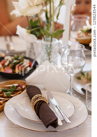 Elegant Wedding Table Setting with White Lillies and Brown Napkins. Formal Dining Scene. Luxury Celebration. Close-up Shot. Soft Lighting 129828544