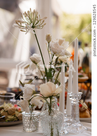 Elegant Wedding Table Setting with White Flowers, Candles, and Crystal Vases: Romantic Dinner Reception, Fine Dining, Luxurious Decor. Elegant Wedding Table Setting with White Flowers, Candles, and Crystal Vases: Romantic Dinner Reception, Fine Dining, Luxurious Decor. 129828545