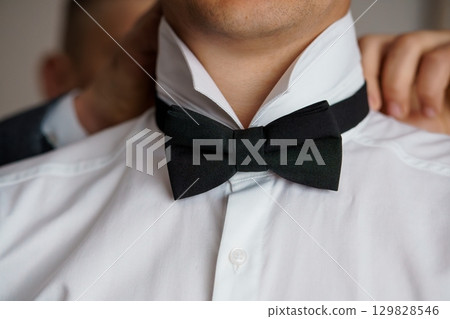 Stylish Man Adjusting Black Bow Tie, Groom Preparation, Formal Attire, Crisp White Shirt, Close-Up Shot, Elegant Wedding Style, Dark Background Stylish Man Adjusting Black Bow Tie, Groom Preparation, Formal Attire, Crisp White Shirt, Close-Up Shot, Elegant Wedding Style, Dark Background 129828546