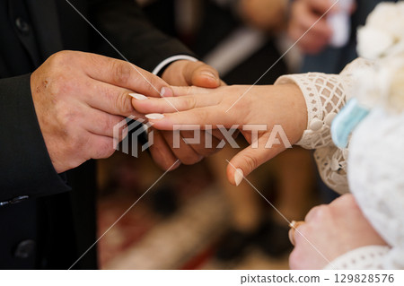 Wedding Ceremony Ring Exchange: Close-up on Hands Placing Wedding Band, Traditional Celebration, Black Suit, White Gown, Formal Setting. 129828576