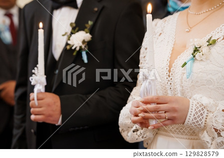 Orthodox Wedding Ceremony with Lit Candles: A Romantic Close-Up of Bride and Groom's Hands Holding Candles, White Lace, Traditional Attire Orthodox Wedding Ceremony with Lit Candles: A Romantic Close-Up of Bride and Groom's Hands Holding Candles, White Lace, Traditional Attire 129828579