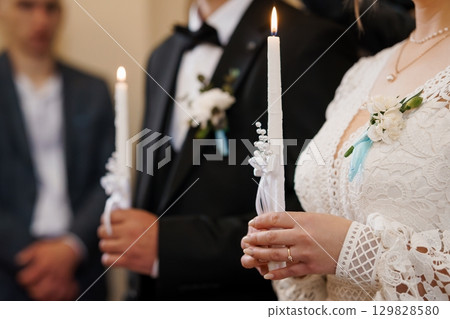 Orthodox Wedding Candle Lighting Ceremony: Groom and Bride Holding Lit Candles in Lace Gown, Dark Suit, Spiritual Ritual. Orthodox Wedding Candle Lighting Ceremony: Groom and Bride Holding Lit Candles in Lace Gown, Dark Suit, Spiritual Ritual. 129828580