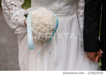 Romantic Wedding Bouquet Held by Bride: Ivory Blossoms, Light Blue Ribbon, Lace Dress and Groom's Hand, Outdoor Ceremony, Elegant Style. Romantic Wedding Bouquet Held by Bride: Ivory Blossoms, Light Blue Ribbon, Lace Dress and Groom's Hand, Outdoor Ceremony, Elegant Style. 129828585