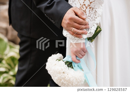 Wedding Hands Close-Up: Groom Holding Bride's Bouquet, Symbolic Love, White Flowers, Elegant Lace Dress, Formal Attire, Outdoor Celebration. 129828587