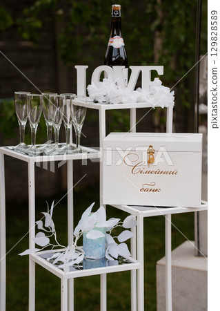 Elegant Wedding Champagne Setup with "LOVE" Sign: Sparkling Toast Celebration, Flutes, Gift Box, White Metal Stand, Lush Greenery. 129828589