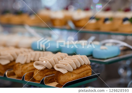 Elegant Pastry Display Featuring Cream Puffs and Macarons on Tiered Trays with Soft Lighting, Selective Focus, Golden Brown and Blue Hues. Elegant Pastry Display Featuring Cream Puffs and Macarons on Tiered Trays with Soft Lighting, Selective Focus, Golden Brown and Blue Hues. 129828593