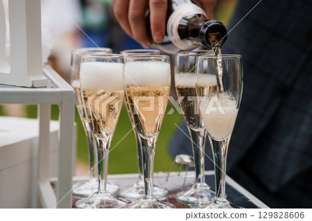 Champagne Pouring Ceremony. Sparkling Wine Toast at Elegant Outdoor Wedding Celebration with Glasses, Tray, Blurred Guests. Champagne Pouring Ceremony. Sparkling Wine Toast at Elegant Outdoor Wedding Celebration with Glasses, Tray, Blurred Guests. 129828606