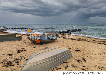 Grey sky looms above rough sea, boats struggle near wet beach. 129828642