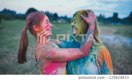 Two female school friends hug each other, covered in colorful powder paint, enjoying the holi festival in a field at sunset Two female school friends hug each other, covered in colorful powder paint, enjoying the holi festival in a field at sunset 129828653