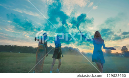 Three school friends enjoying a vibrant sunset while playing with blue smoke bombs in a field, creating a colorful and memorable scene Three school friends enjoying a vibrant sunset while playing with blue smoke bombs in a field, creating a colorful and memorable scene 129828660