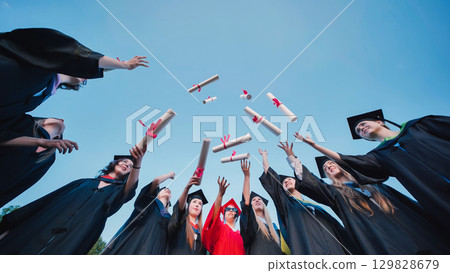 Group of students wearing graduation gowns and caps, joyfully throwing diplomas into the air against a clear blue sky, celebrating their academic achievement Group of students wearing graduation gowns and caps, joyfully throwing diplomas into the air against a clear blue sky, celebrating their academic achievement 129828679