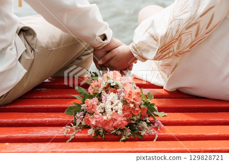 Romantic Wedding Ceremony Hands Holding Bouquet on Red Boat Lake Shore, Traditional Ukrainian Style Embroidery, Warm Pastel Colors. Romantic Wedding Ceremony Hands Holding Bouquet on Red Boat Lake Shore, Traditional Ukrainian Style Embroidery, Warm Pastel Colors. 129828721