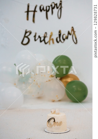 First Birthday Celebration with Balloons and Cake, Gold Lettering and Green Decor, Festive Party Scene, White Backdrop, One Year Old Event. 129828731