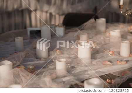Romantic Indoor Scene with Candlelit Table Setting and Rose Petals, Soft White Fabrics, and Intimate Ambiance for Celebration. Romantic Indoor Scene with Candlelit Table Setting and Rose Petals, Soft White Fabrics, and Intimate Ambiance for Celebration. 129828735