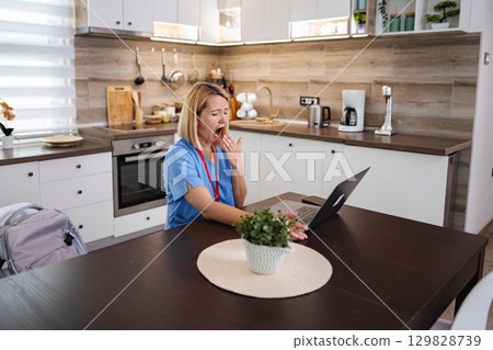 Exhausted Female Doctor Yawning While Working on Laptop in Kitchen Exhausted Female Doctor Yawning While Working on Laptop in Kitchen 129828739