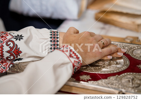 Hands Praying on Holy Book with Embroidered Sleeves, Orthodox Christian Tradition, Golden Cover, Red Textile, Spiritual Devotion, Close-Up 129828752