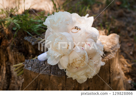 Romantic Wedding Rings Resting on White Peony Bouquet Placed on Tree Stump in Forest, Golden Bands, Soft Lighting, Love, Marriage, Engagement 129828866