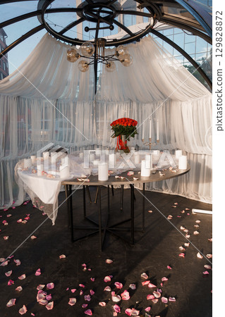 Romantic Dinner Setup in Glass Dome with Red Roses, Candles and Draped White Fabric. Intimate Evening Mood, Dark Wood Table, Warm Lighting. 129828872