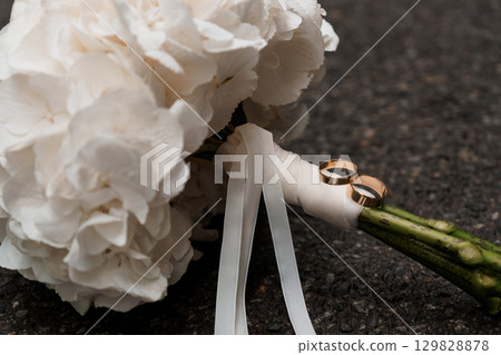 Wedding Rings on Bouquet: Romantic Bridal Close-up with White Flowers, Silk Ribbon and Dark Backdrop - Love, Commitment and Nuptial Symbolism 129828878