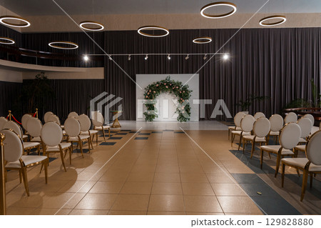 Elegant Wedding Ceremony Setup in a Modern Ballroom with Dramatic Curtains, Floral Arch, Golden Chairs and Ambient Lighting 129828880