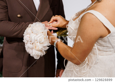 Romantic Wedding Ceremony Exchange Rings Close-Up. Brown Suit, White Gown, Peony Bouquet. Symbolic Love Gesture 129828899