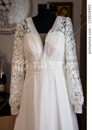 Elegant White Lace Wedding Dress on Mannequin. Boho Style Gown with Long Sleeves, Detailed Embroidery, Soft Fabric, Rustic Interior Setting 129828905