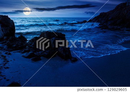 landscape by the sea at night in summer. beautiful beach with rocks washed by calm waves in full moon light. view to horizon under dark sky with clouds. scenic travel destination for leisure and relax 129829605