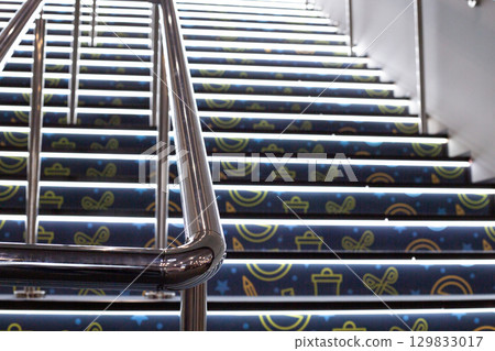 Close up of metal handrail of illuminated staircase, with glowing steps in background. The sleek modern design of handrail contrasts with the vibrant light of the steps 129833017